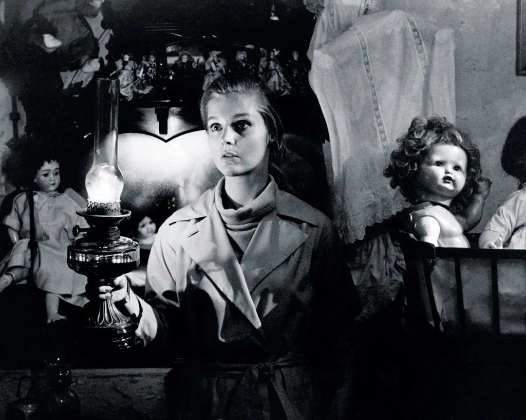 black and white still from Bunny Lake Is Missing. Image shows a white woman in low lighting, surrounded by creepy dolls.
